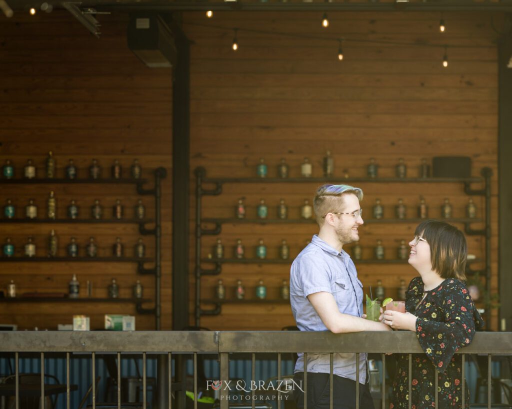 Engaged couple holding colorful drinks at Bluebird Distilling railing in Phoenixville perfect spot for Phoenixville engagement sessions