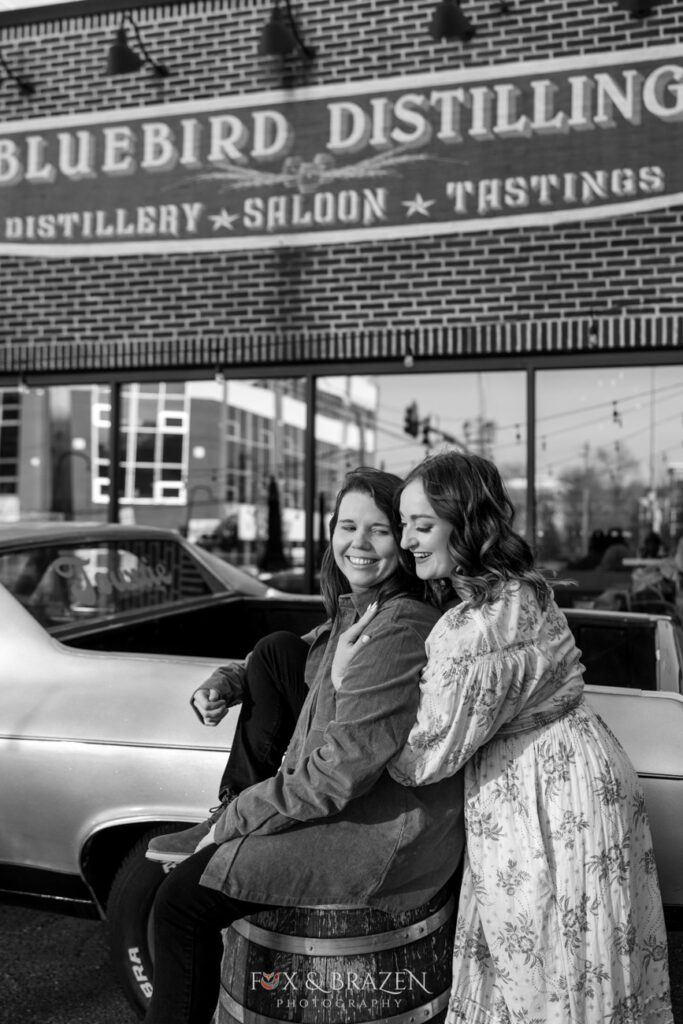 Black-and-white photo of engaged couple near the El Camino outside Bluebird Distilling during their engagement session.