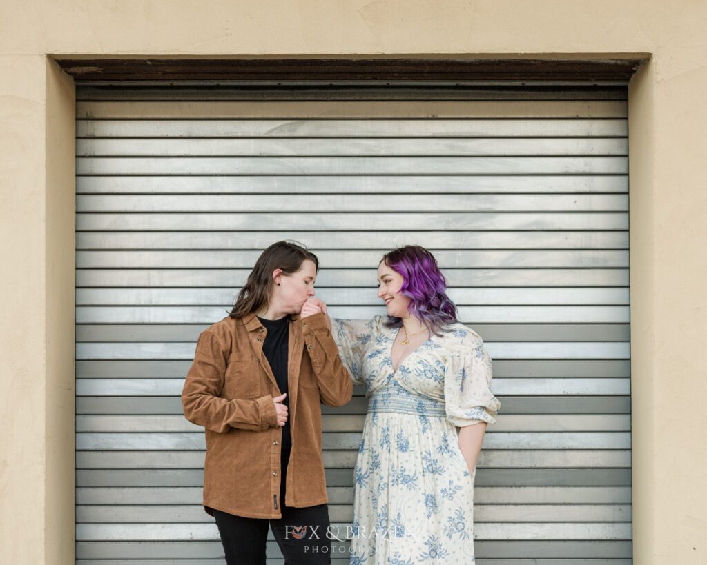 Engaged couple sharing a kiss on the hand near a silver loading dock door for their Downtown Phoenixville engagement adventure