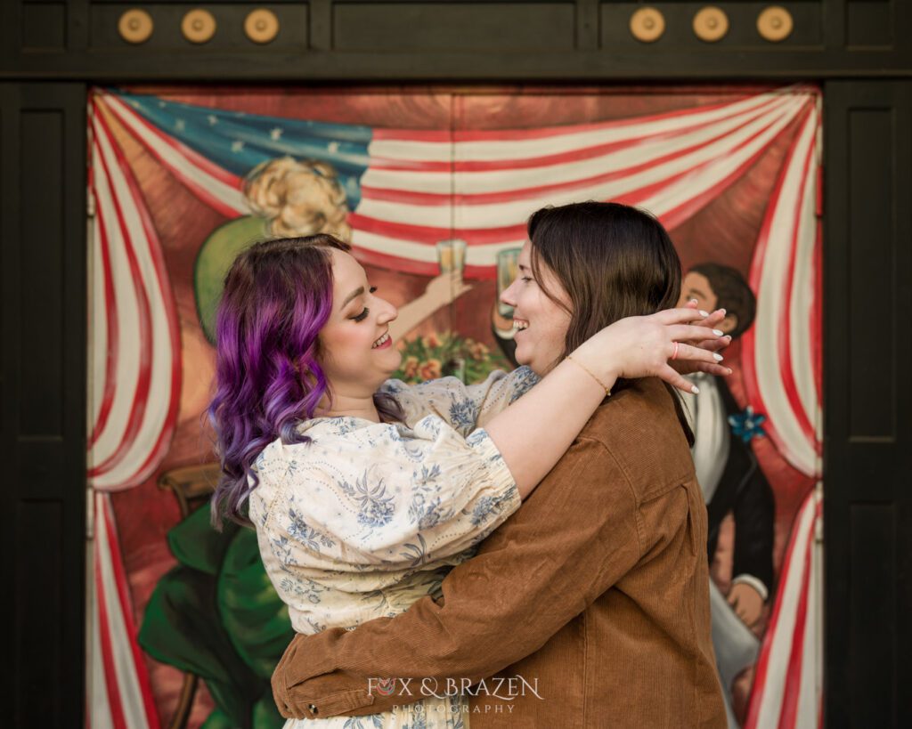 Engaged couple looking at each other and embracing in front of a mural on Bridge Street during their engagement session.