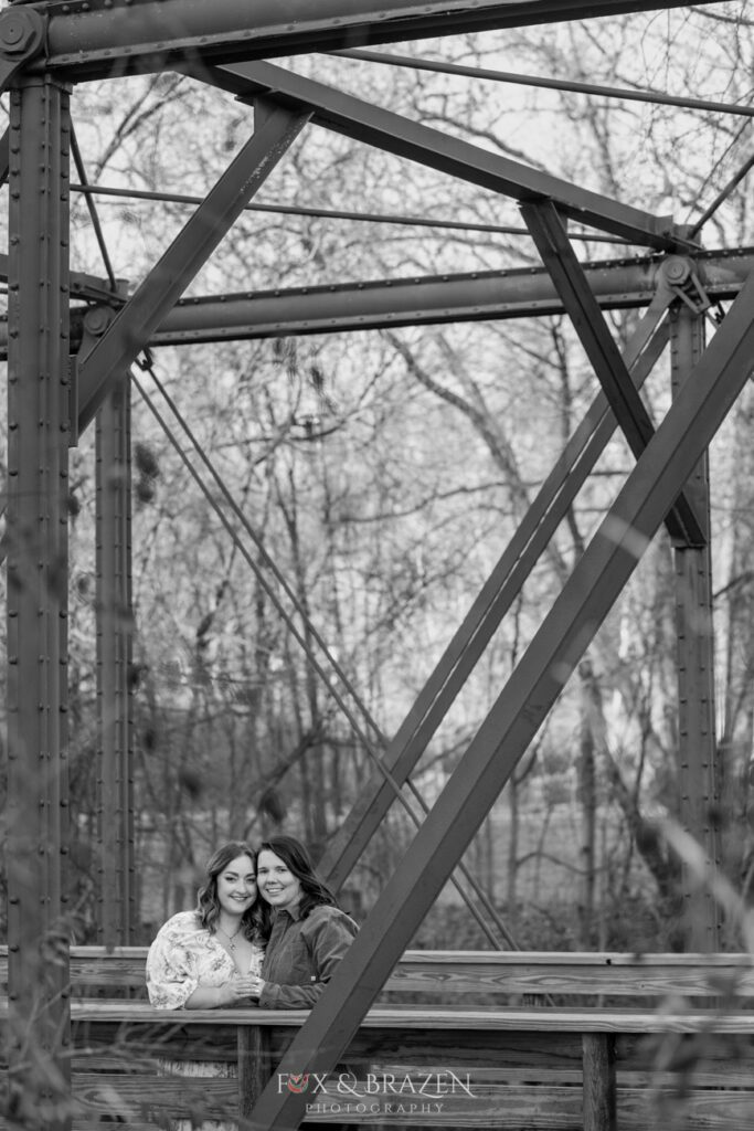 Engaged couple looks at the camera while standing on a bridge in black and white.
