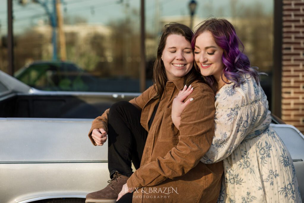 Engaged couple snuggling together near an El Camino outside Bluebird Distilling.