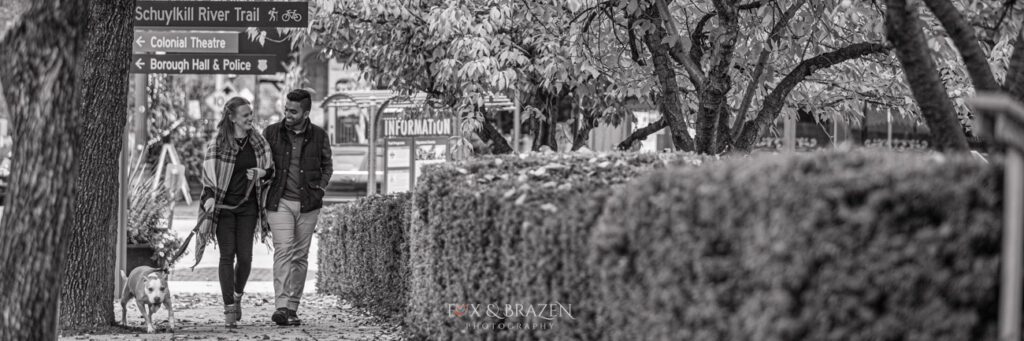 Engaged couple walking dog on Main Street in Phoenixville the perfect town for Phoenixville engagement sessions