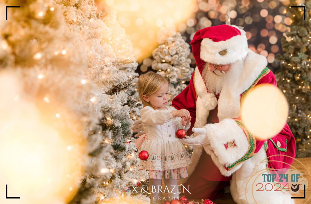Santa helps a little girl decorate a Christmas tree on a festive set.