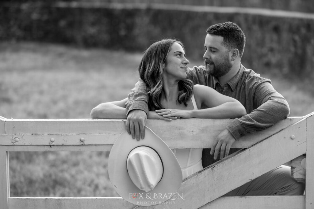 Black-and-white photo of a couple standing by a fence, gazing at each other lovingly.