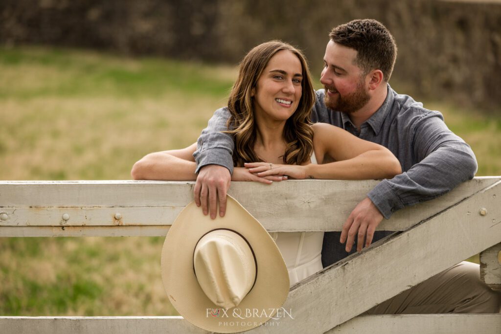 Couple leaning on a fence with a cowboy hat during engagement photos.