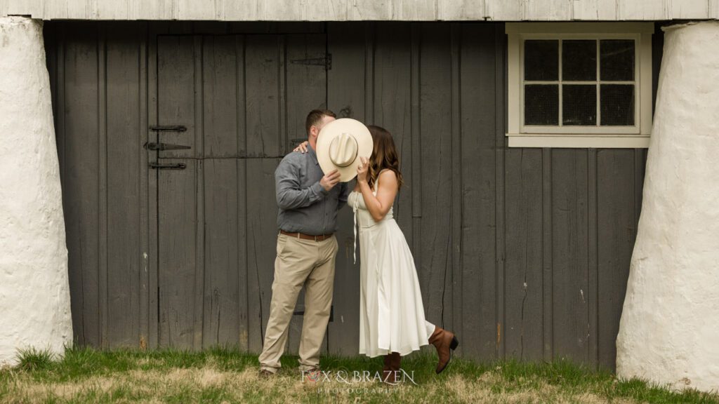 Couple kissing behind a cowboy hat during engagement photography.