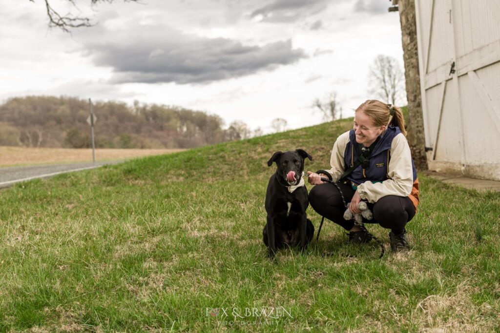 Woman kneels with black dog as the handler at a Valley Forge Engagement Photography Session in the spring.