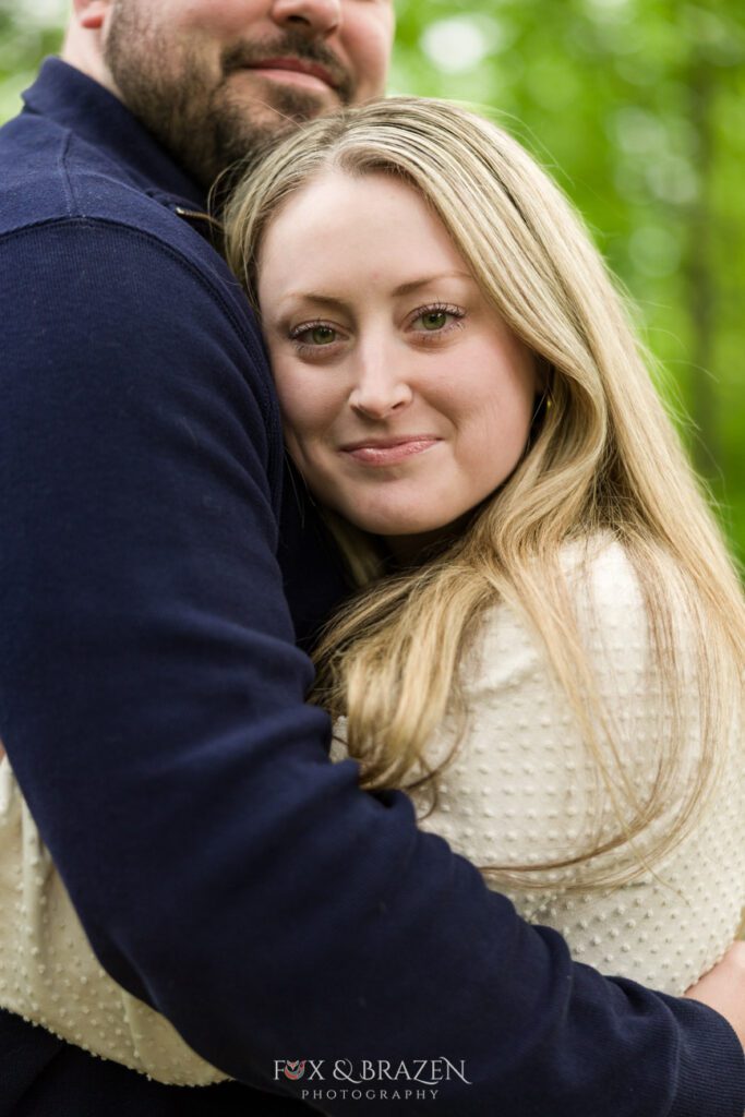 Woman looking content into the camera while being embraced by her fiancé during Valley Forge session.