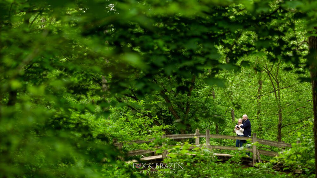 Couple standing on a bridge surrounded by a canopy of green trees at Valley Forge.