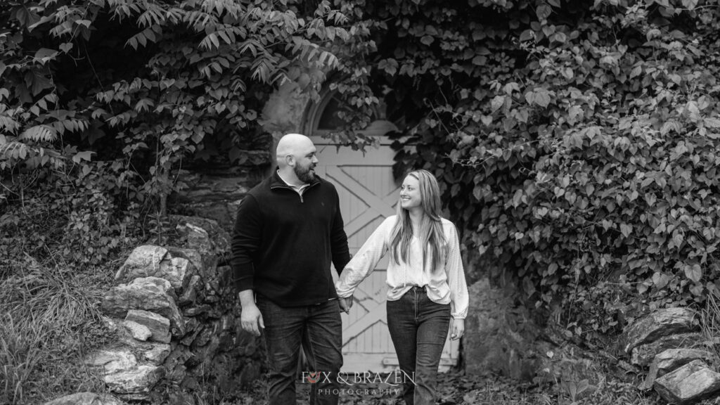 Black and white photo of engaged couple walking with autumn foliage in the background at Valley Forge.
