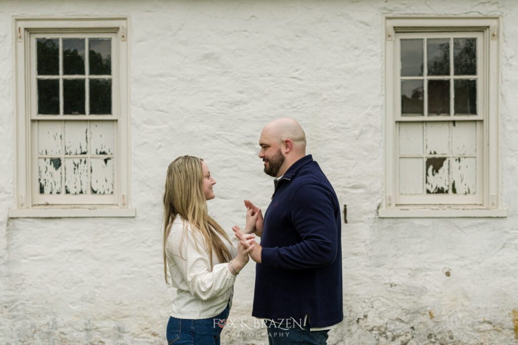 Couple standing in front of a white wall, holding hands and looking at each other during engagement session.