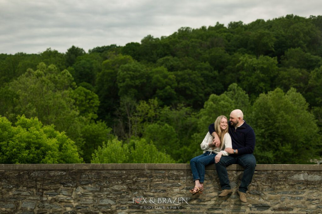Engaged couple sitting together on a stone wall under a dramatic dark sky at Valley Forge.