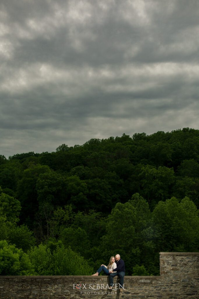 Couple sitting on a stone wall under a stormy sky during their Valley Forge engagement session.