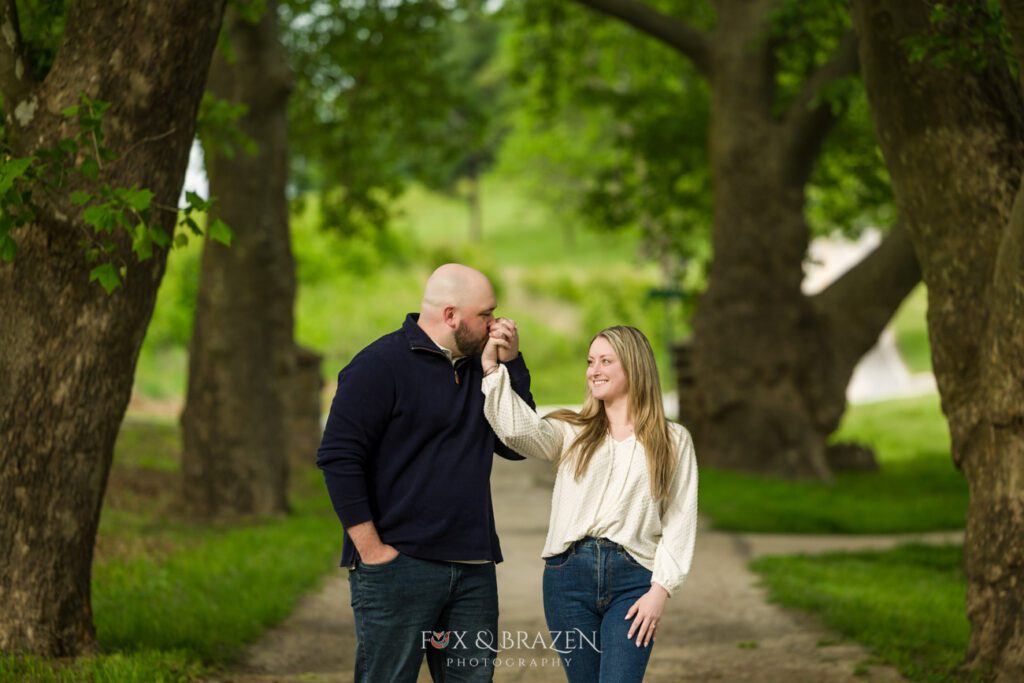 Man kissing his fiancée’s hand as she smiles during their Valley Forge engagement session.