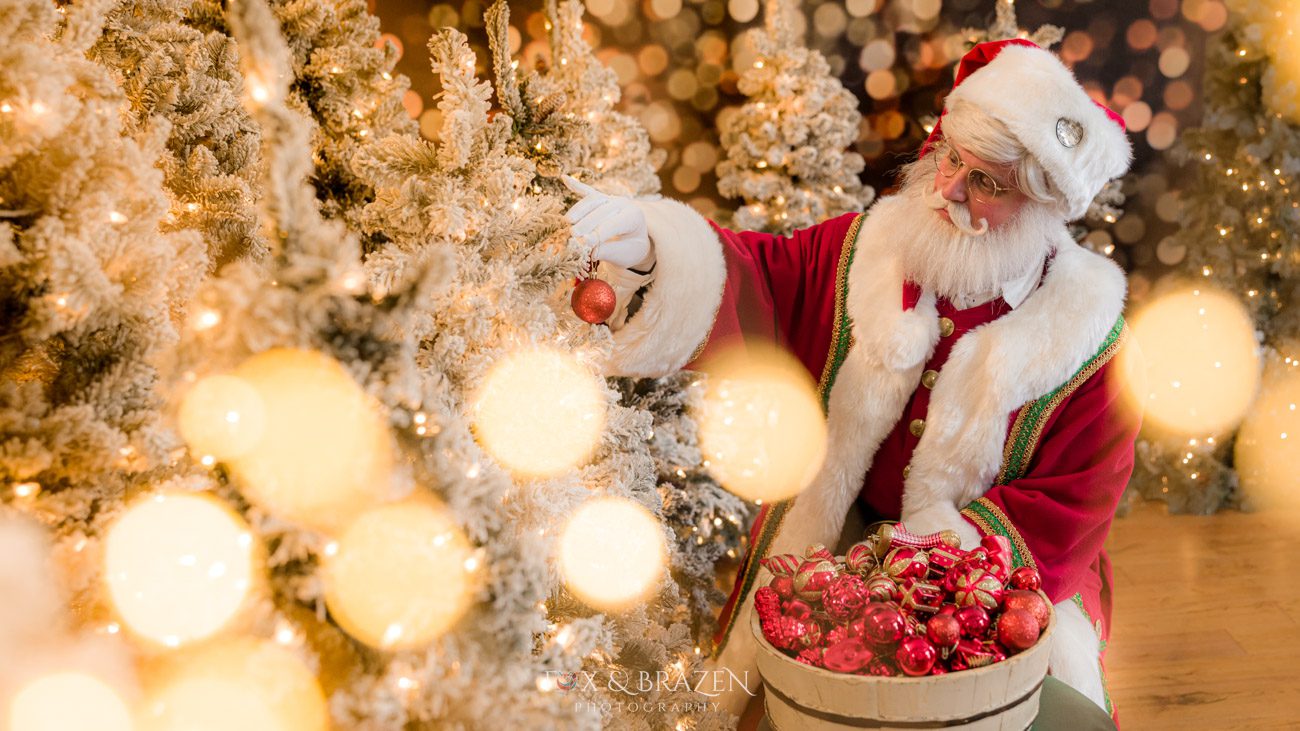 Santa Claus surrounded by twinkling lights and red ornaments while decorating Christmas trees.